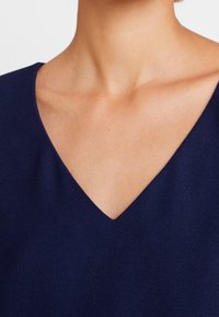 Mörk marinblå V-ringad topp med en slät textur och minimalistisk design, som visar en subtil drapering runt halsen och axlarna.