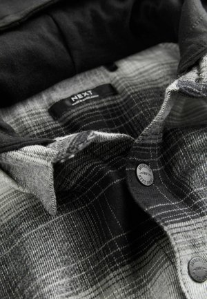 Chemise en flanelle avec un motif à carreaux noirs et gris, une texture douce, une fermeture à boutons et une étiquette portant la mention "NEXT".