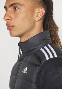 Crni podstavljeni bezrukavnik s visokim ovratnikom, s bijelim Adidas logotipom i tri bijele pruge na ramenu. Glatka tekstura.