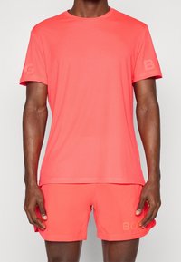 Světle korálové sportovní tričko a šortky, vyrobené z hladkého materiálu s texturovaným vzorem, obsahující loga na rukávech a šortkách.