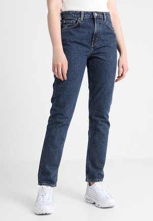 Personne portant un jean taille haute bleu foncé et des baskets blanches, debout devant un fond clair uni.