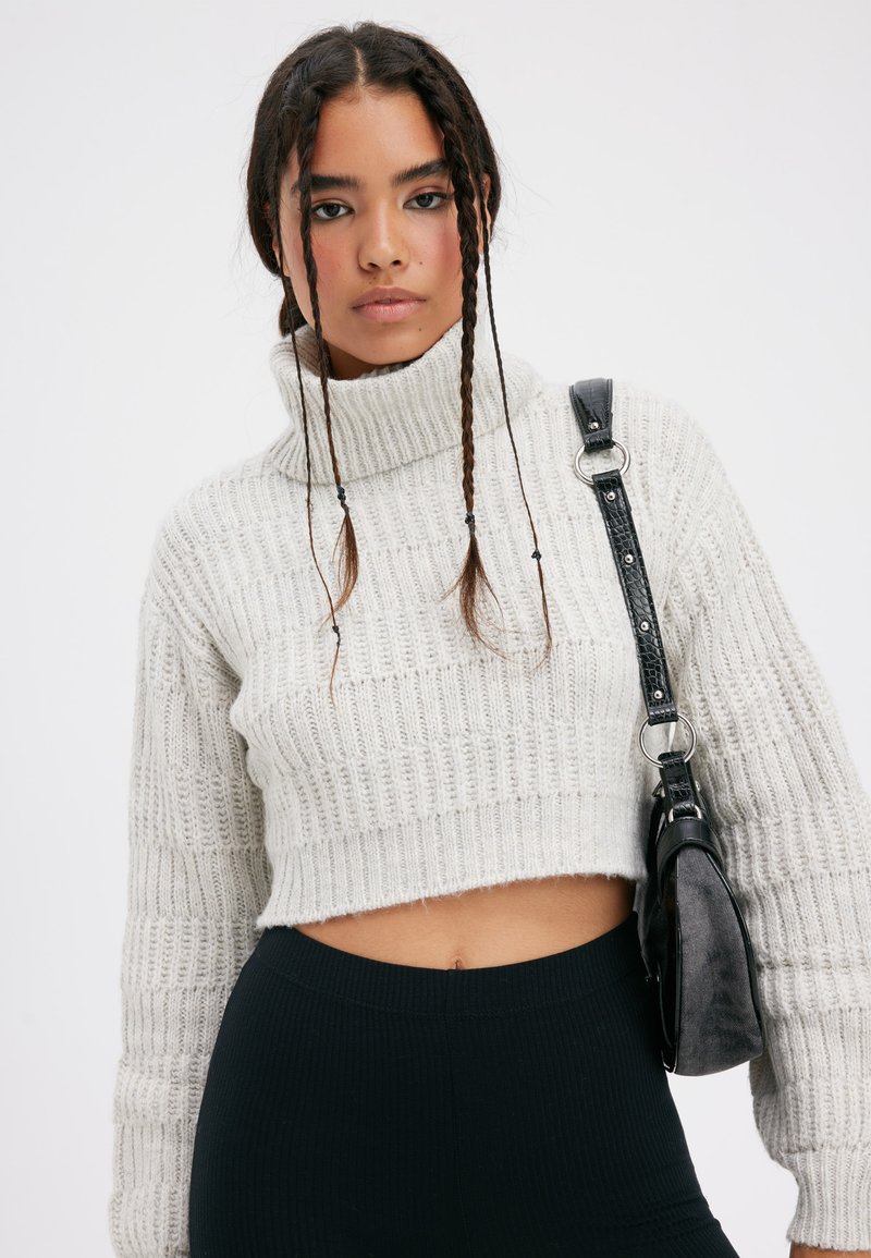 Pull court style crochet en gris clair, avec un col montant et une texture côtelée. Associé à un sac d'épaule noir texturé.