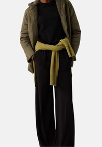 Prošívaná olivově zelená bunda s límcem z beránčí kožešiny, černá košile, černé kalhoty s širokými nohavicemi a žluto-proužkovaná pletená šála uvázaná v pase.