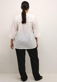 Hvid bluse med polka prikker, puffede ærmer og rund kant, parret med sorte lige bukser i en glat stofstruktur.