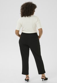 Une femme aux cheveux bouclés porte un haut blanc à manches courtes, un pantalon noir taille haute et des sandales à talons noires, se tenant avec les mains dans les poches.