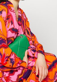 Groene leren clutch met een quilted patroon en een zilveren ketting, vastgehouden tegen een levendig, patroonrijke jurk met lange mouwen in roze en oranje.