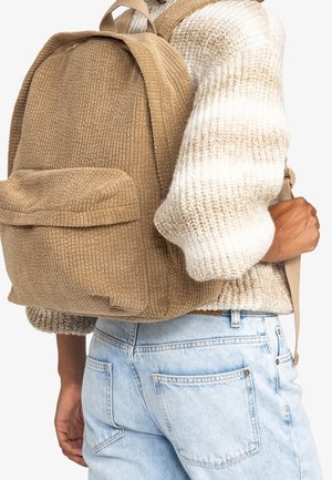 Osoba nosząca beżowy plecak z prążkowanej tkaniny oraz gruby, kremowo-brązowy sweter w paski wykonany na drutach, a do tego jasnoniebieskie dżinsy.
