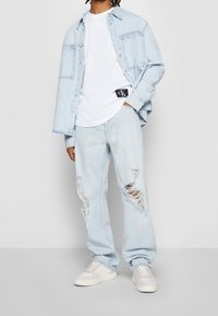 Jasnoniebieska koszula dżinsowa na białym T-shircie, w zestawieniu z jasnoniebieskimi jeansami z przetarciami i białymi sneakersami. Zauważalne przetarcia z przodu spodni.