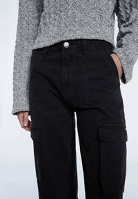Pantalon cargo noir avec poches latérales, taille haute, tissu robuste et fermeture à bouton argenté. Porté avec un pull gris texturé.