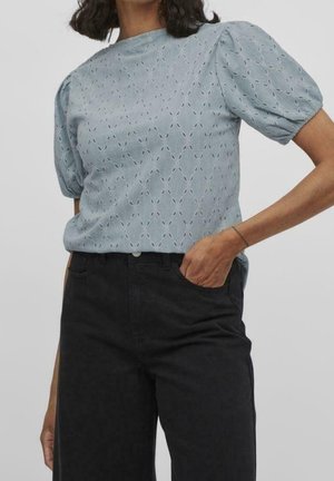 Jasnoniebieska bluzka z krótkim rękawem i bufkami, z teksturowanym wzorem i wysokim dekoltem. W zestawie z czarnymi spodniami.