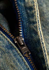 Nahaufnahme eines Denim-Stoffes mit einem goldenen Reißverschluss und einem schwarzen Zugriegel, der strukturierte Blautöne und sichtbare Nähdetails zeigt.