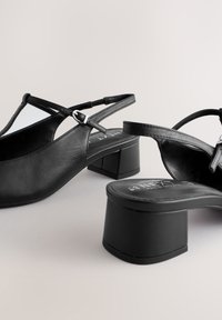 Escarpins slingback en cuir noir avec talons blocs et boucles argentées réglables, présentés sur un fond neutre.