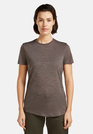 Kvinde med kort mørkt hår iført en tætsiddende kortærmet brun meleret t-shirt og olivengrønne bukser, stående mod en ensfarvet baggrund.