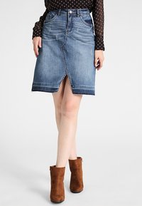 Denim kynähame, jossa on rikottu helma, etuhalkio ja viisi taskua. Yhdistetty ruskeiden mokkanahkasaappaiden ja mustan pilkullisen paidan kanssa.