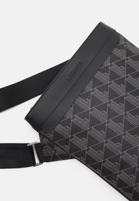 Bolso cruzado de tela negra con un diseño geométrico, detalles lisos en negro, correa ajustable y un logo sutil.