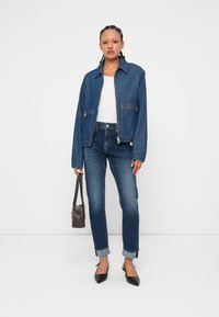 Donna in piedi che indossa una maglietta bianca, una giacca di jeans blu, jeans arrotolati, scarpe nere a punta e tiene in mano una piccola borsa marrone.