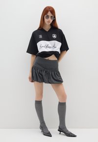 Camisa deportiva de gran tamaño en blanco y negro, falda corta gris con dobladillo con volantes, calcetas gris hasta la rodilla y tacones negros puntiagudos con studded.