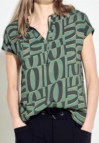 Blusa de manga corta, con un patrón en verde y negro; presenta un escote en V, detalle de botones y un corte relajado con una tela texturizada.