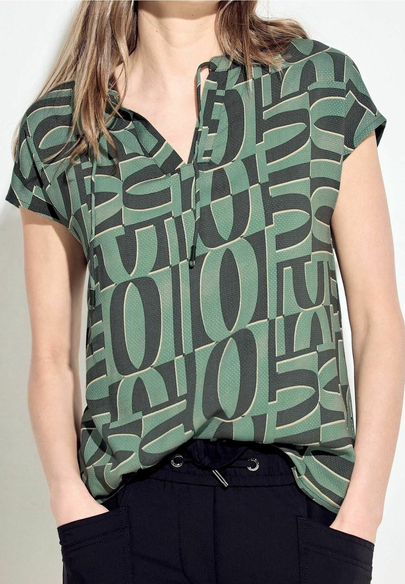 Blusa de manga corta, con un patrón en verde y negro; presenta un escote en V, detalle de botones y un corte relajado con una tela texturizada.