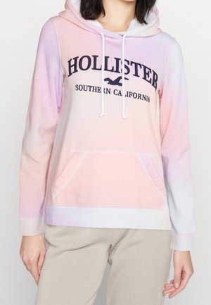 Mujer con una sudadera con capucha Hollister de degradado en tonos rosa pastel y púrpura con el texto "Southern California" y pantalones beige, con las manos relajadas a los lados.
