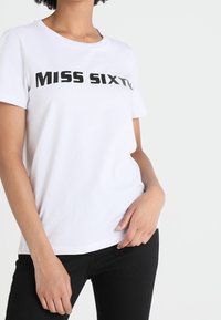 Vit kortärmad T-shirt med texten "MISS SIXTY" bärs av en person med handen vilande nära fickan, i kombination med svarta byxor.