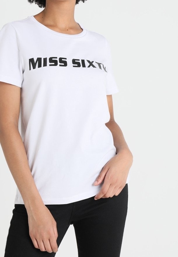 Vit kortärmad T-shirt med texten "MISS SIXTY" bärs av en person med handen vilande nära fickan, i kombination med svarta byxor.