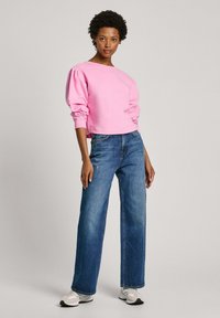 Rozā īsā kapucēta džemperis ar pūkainām piedurknēm, savienots ar augsto vidukli ziliem džinsiem. Modelis valkā baltas sporta kurpes. Vienkāršs, minimālistisks stils.