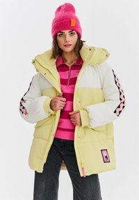 Bunda s výplní v žltej a bielej farbe s rebrovanou textúrou. Obsahuje ružové akcenty a grafický nášivku. Modelka nosí ružový čiapku a pruhovaný top.