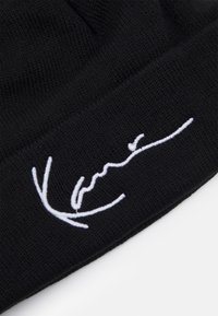 Svart strikket lue med brett, med hvit brodert signaturdetalj over forsiden. Myk, teksturert stoff.