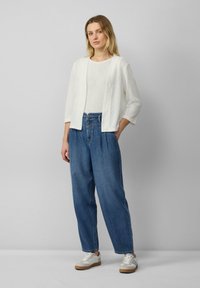 Weißer, strukturierter Cardigan über einem weißen Oberteil, kombiniert mit lockeren blauen Jeans. Die Person trägt leichte Sneaker mit beigen Akzenten.