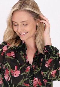 Bluză neagră transparentă cu model floral roșu, detalii cu nasturi și mâneci lungi. Are un guler clasic și o ușoară adunare la manșetă.