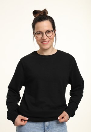 Glunderende vrouw met een bril en een haarbun, gekleed in een zwarte sweater en lichtblauwe jeans, die de onderkant van haar sweater vasthoudt tegen een effen achtergrond.