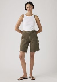 Γυναίκα που φοράει λευκό αμάνικο τοπ, χακί σορτς με ξέφτια και μεγάλες τσέπες, και μαύρες σαγιονάρες, στέκεται με τα χέρια πίσω από την πλάτη.