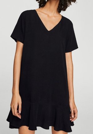 Mujer con un vestido negro de manga corta, escote en V y bajo con volantes, de pie frente a un fondo liso de color gris claro.