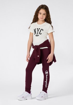 Camiseta blanca con gráfico "NYC 19", combinada con leggings burdeos que presentan el logo de "Champion". Zapatillas blancas con acentos morados completan el conjunto.