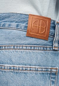 Τζιν παντελόνι από ανοιχτό μπλε τζιν με πορτοκαλί ραφές, καφέ δερμάτινο λογότυπο με ανάγλυφα γράμματα στη ζώνη μέσης και λευκό ύφασμα που προεξέχει.