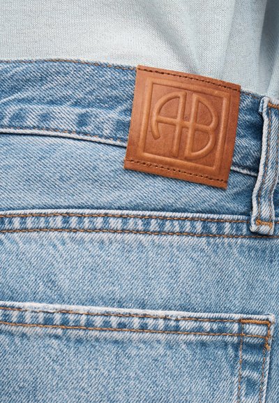 Lyseblå denimjeans med oransje søm, en brun lærmerke med preget skrift på linningen, og et hvitt stoff som er stukket inn.