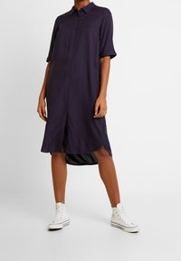 Mörkblå skjortklänning med avslappnad passform, korta ärmar, en hög-låg fåll och en mjuk textur, tillsammans med vita hög-top sneakers.