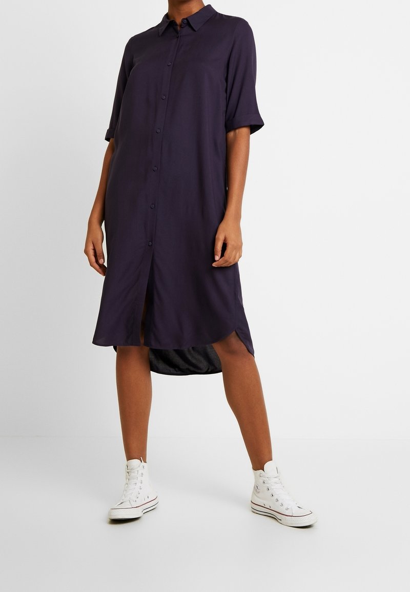 Mörkblå skjortklänning med avslappnad passform, korta ärmar, en hög-låg fåll och en mjuk textur, tillsammans med vita hög-top sneakers.