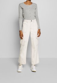 Szary długi kardigan z długim rękawem i zapięciem na guziki, w połączeniu z kremowymi spodniami cargo z przednimi kieszeniami i luźnym krojem, uzupełnionymi białymi sneakersami.