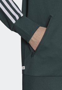 Dark green athletic jacket with white stripes, featuring a zippered pocket on the side and textured cuffs for a secure fit.