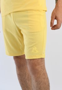 Pantalones cortos de algodón amarillos con un diseño ajustado, que presentan un logotipo en el lado inferior izquierdo y costuras decorativas alrededor del dobladillo.