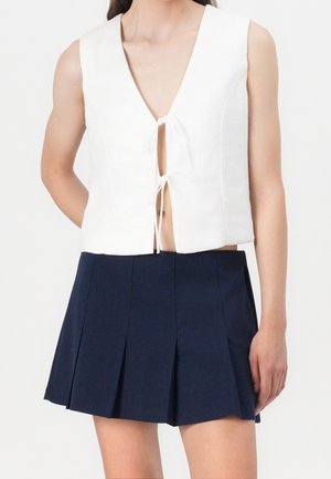 Haut blanc sans manches avec liens à l'avant, associé à une mini-jupe plissée taille haute bleu marine sur une personne debout.
