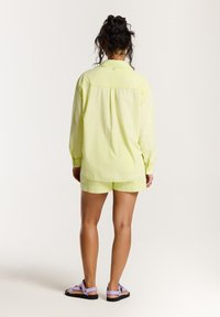 Lichtgele oversized shirt met lange mouwen, gecombineerd met bijpassende shorts. De outfit bevat paarse sandalen met verstelbare banden.
