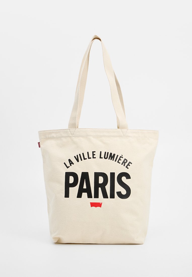 Borsa tote in canvas beige con testo grafico nero che recita "LA VILLE LUMIÈRE PARIS" e un piccolo logo rosso in basso. Manici lunghi.