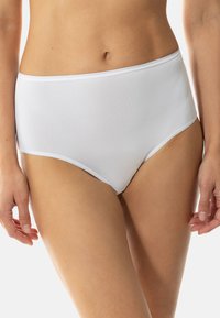 Frau in schlichten weißen Taillenhosen, die mit entspannten Händen an den Seiten vor einem weißen Hintergrund steht.