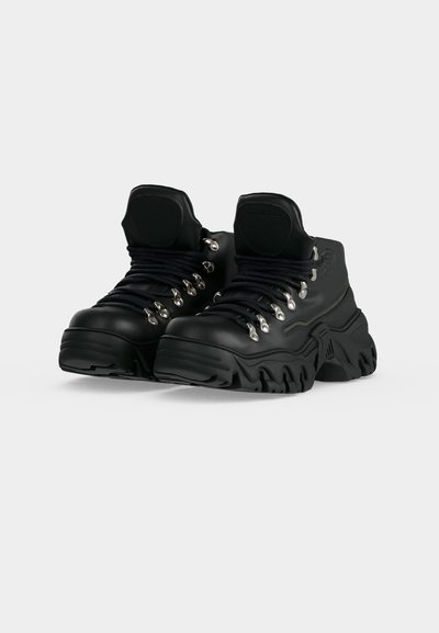 Μαύρα χοντρά μπότες πλατφόρμας με παχιά υφασμένα πέλματα και μαύρα κορδόνια περασμένα από ασημένιες τρύπες σε απλό λευκό φόντο.