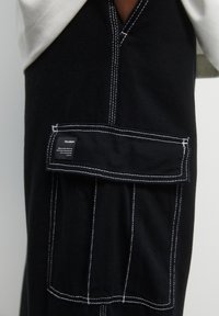 Zwarte cargobroek van stof met witte stiksels, voorzien van een grote voorkantzak en een gemerkt label aan de zijkant.
