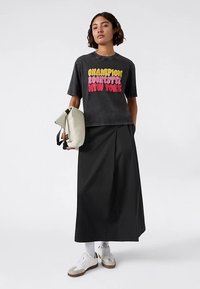 Zwart grafisch T-shirt met de tekst "Champion Rochester New York" in geel en roze, gecombineerd met een zwarte rok en witte sneakers.