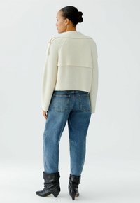 Frau mit einem Dutt, die einen cremefarbenen Strickpullover, eine blaue Jeans und schwarze, lässige Stiefeletten trägt, steht vor einem schlichten weißen Hintergrund.